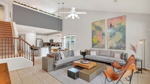 A modern living room with a sofa, coffee table, and chairs, a staircase, and a kitchen in the background.