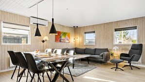 A modern living room with a dining area, featuring a wooden paneled wall, a grey sofa, a dining table with chairs, and pendant lighting.
