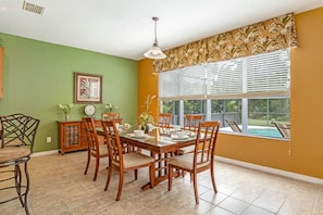 A dining area with a wooden table and chairs, a green wall, a window with blinds, and a view of a swimming pool.