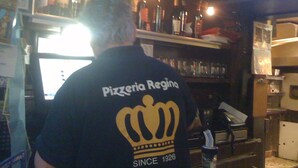A bar with shelves of bottles and glasses, a computer monitor, and a person wearing a 'Pizzeria Regina' t-shirt.