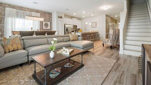 A modern living room with a sectional sofa, a coffee table with magazines, a chandelier, and a staircase leading to the upper floor.