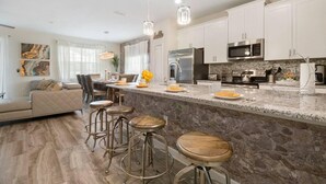 A modern kitchen with white cabinets, stainless steel appliances, and a granite countertop. There are bar stools and a dining area with a sofa and chairs.