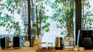 Une table en bois, sur laquelle se trouvent le matériel nécessaire pour préparer du café, des bocaux et un présentoir à menus, est placée près d’une fenêtre donnant sur un espace verdoyant.