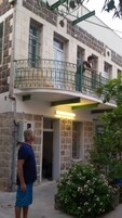 Un edificio de dos pisos con un balcón, fachada de piedra y un pequeño jardín con flores rosadas.