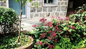 Un bâtiment en pierre doté d’une fenêtre et d’une porte, entouré d’un jardin où poussent des arbustes verts et des fleurs roses.
