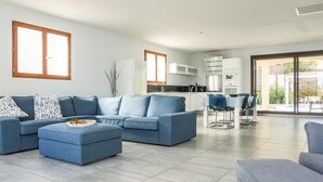 A modern living room with a sectional sofa, a dining area with a table and chairs, and a kitchen with white cabinets and a large window.