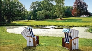 Zwei Korbstühle mit gestreiften Kissen blicken auf einen kleinen Teich in einem Park.