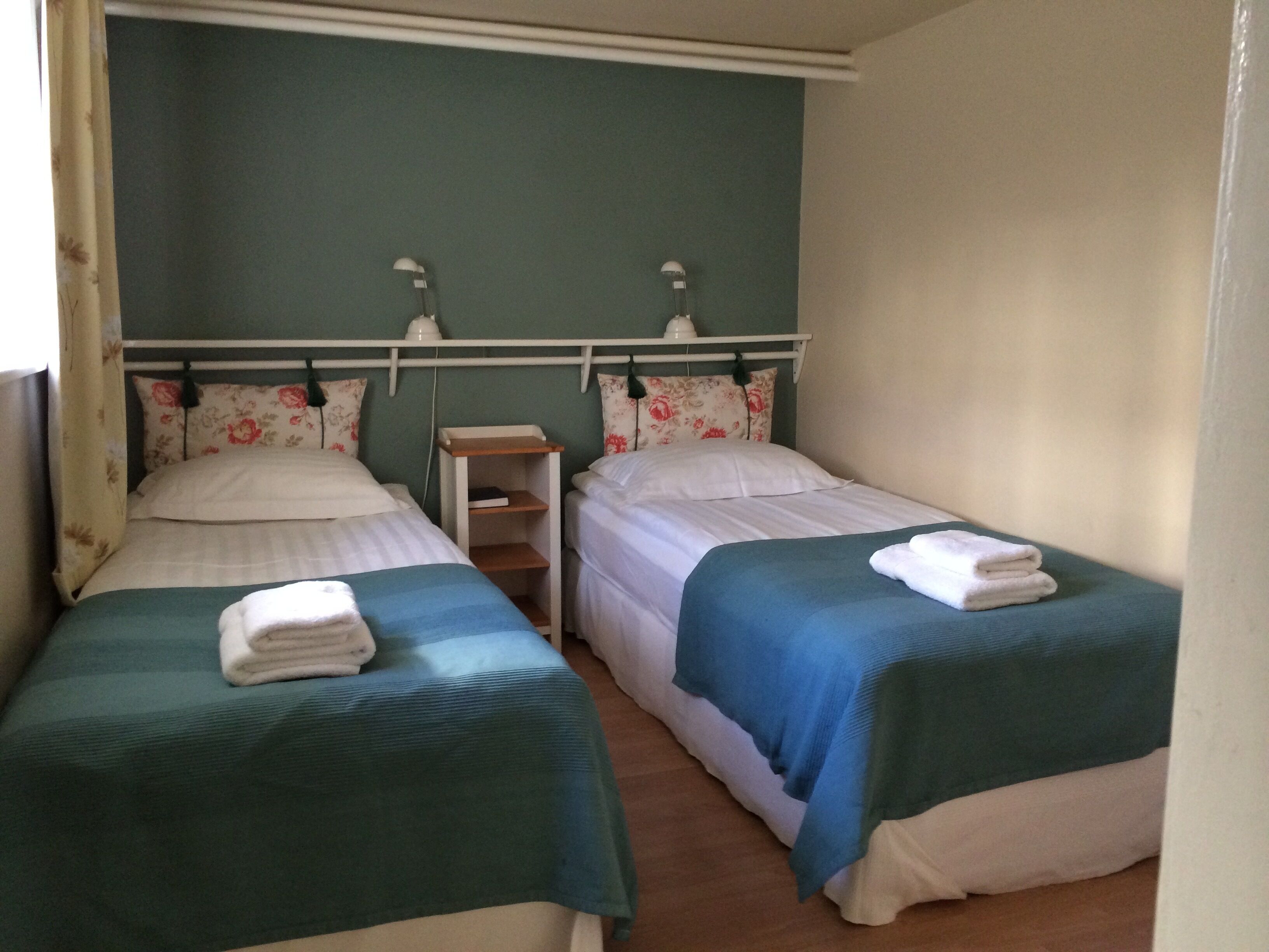 Habitación doble con dos camas individuales, cada una con una colcha verde y almohadas blancas. Hay una mesita de noche con una lámpara entre las camas.