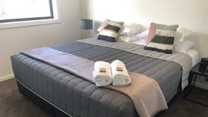 Una cama doble con cubrecolcha gris acolchado, dos toallas enrolladas y una manta beige.