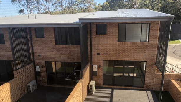 Un moderno edificio de dos plantas con fachada de ladrillo, ventanas amplias y un espacio al aire libre cubierto con patio y zonas verdes.