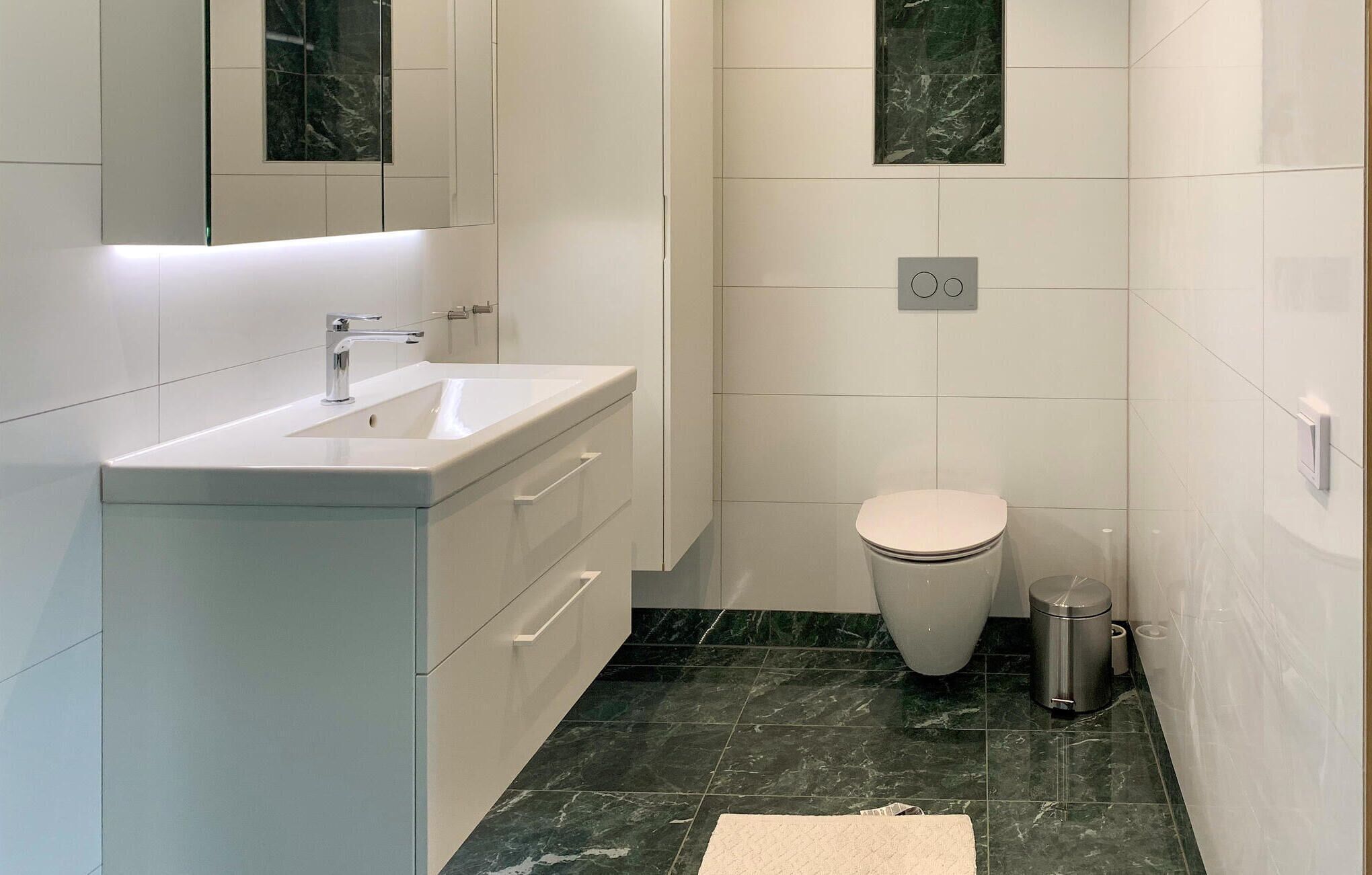 A modern bathroom with a white sink, vanity, and toilet. The floor is tiled with dark marble, and there is a trash can and a towel on the floor.
