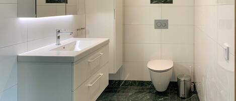 A modern bathroom with a white sink, vanity, and toilet. The floor is tiled with dark marble, and there is a trash can and a towel on the floor.