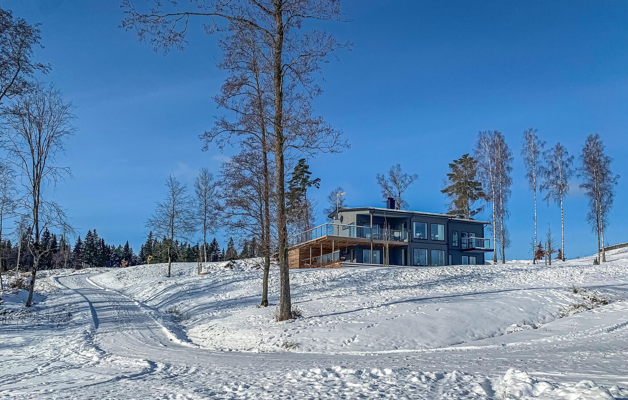 Una cabaña moderna de dos pisos con un balcón, rodeada de suelo cubierto de nieve y árboles escasos.