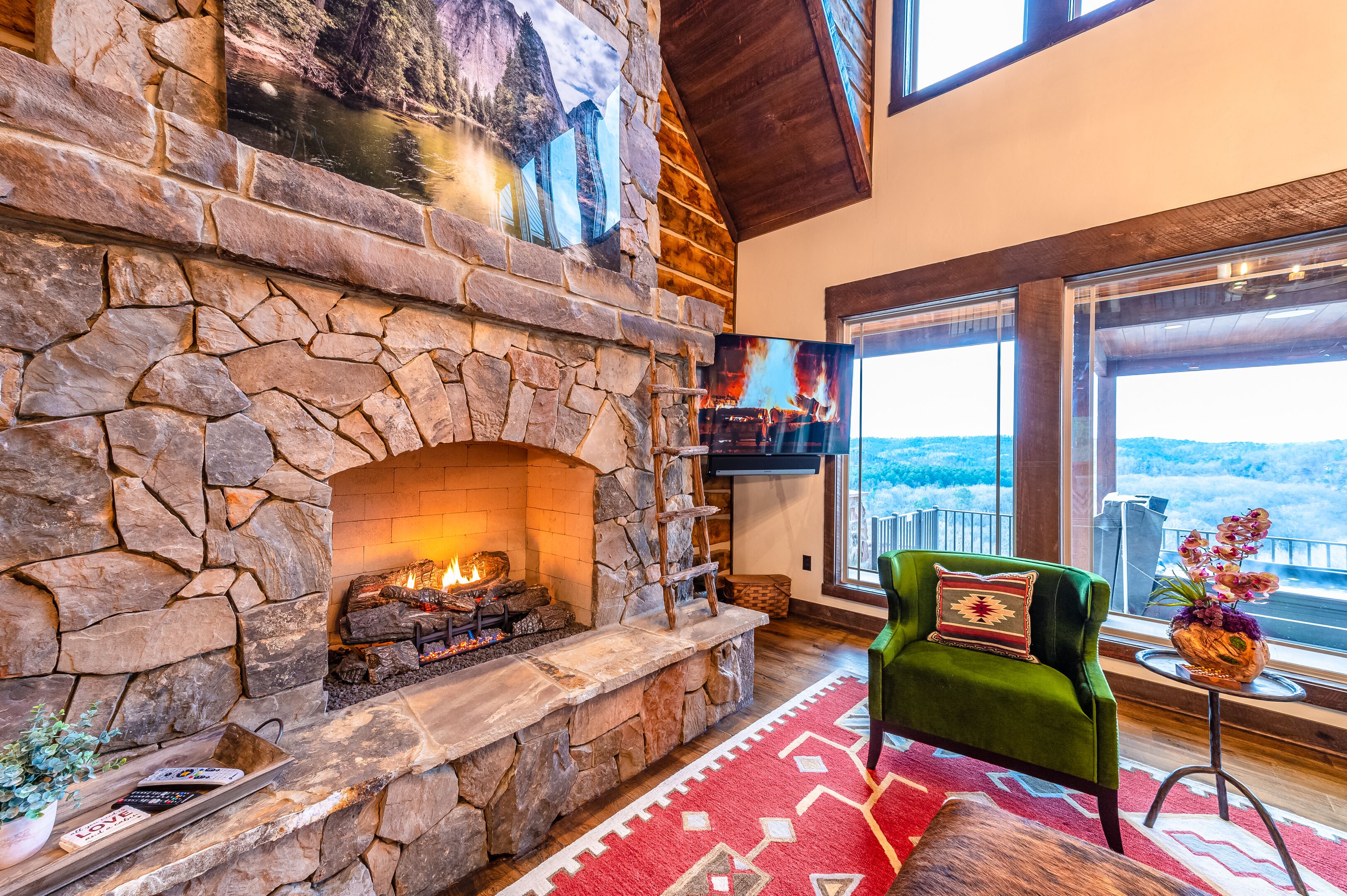 A stone fireplace with a fire, a green armchair with a decorative pillow, and a view of the outdoors through large windows.