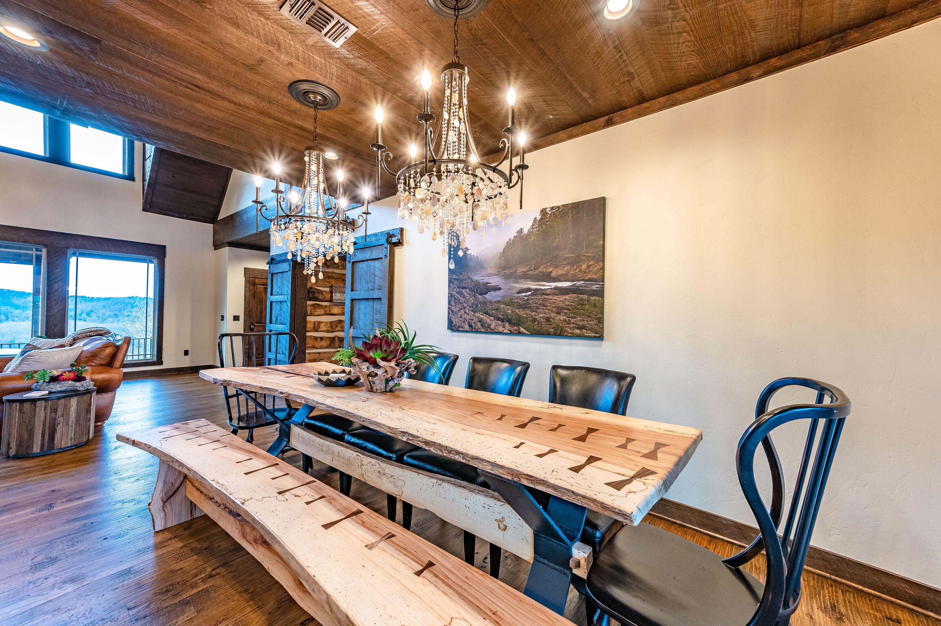 Un comedor espacioso con una larga mesa de madera, bancos y sillas modernas. Un candelabro cuelga del techo, y un cuadro grande decora la pared.
