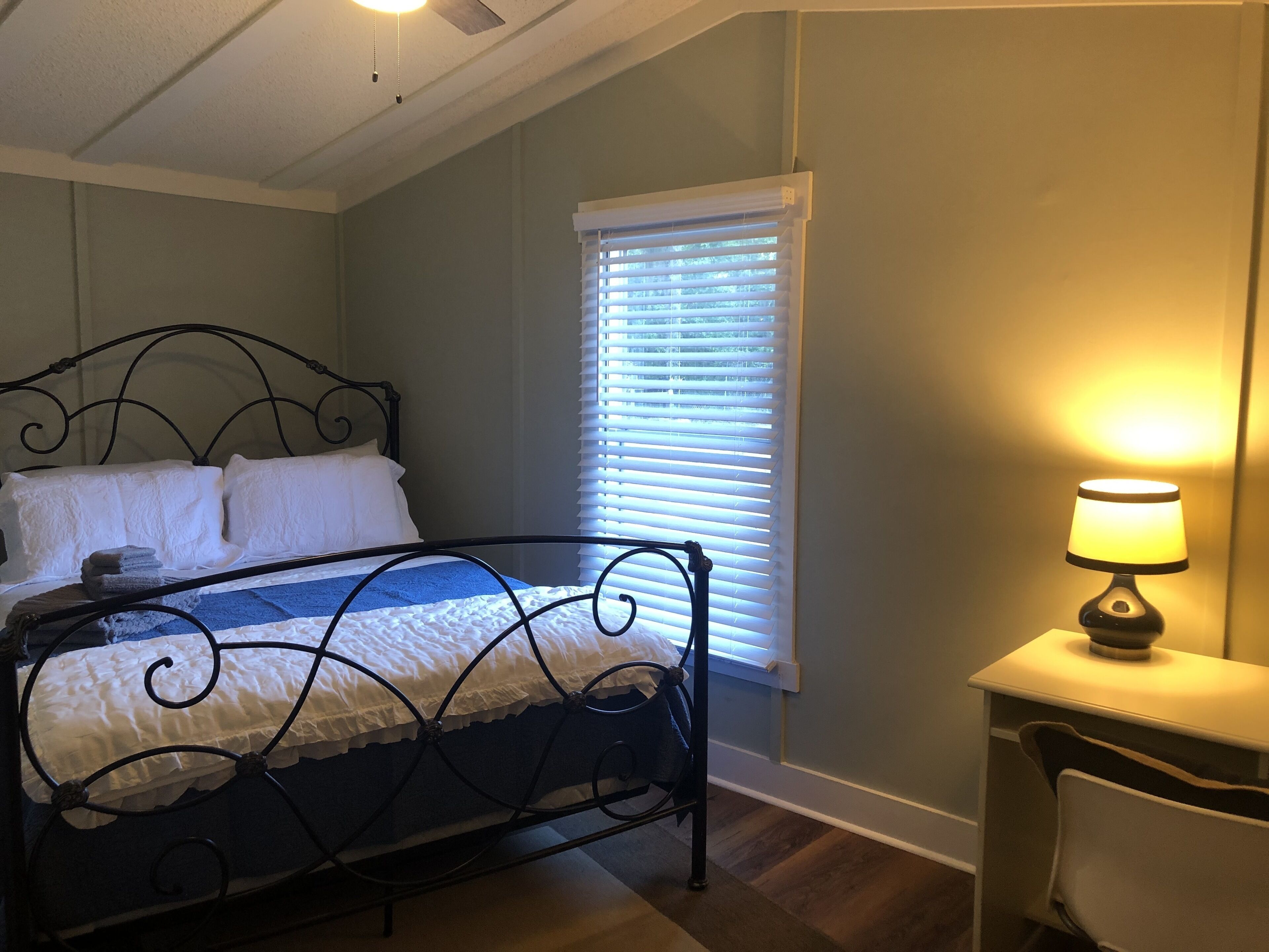 A bedroom with a metal bed frame, a bedside table with a lamp, a window with blinds, and a ceiling fan.