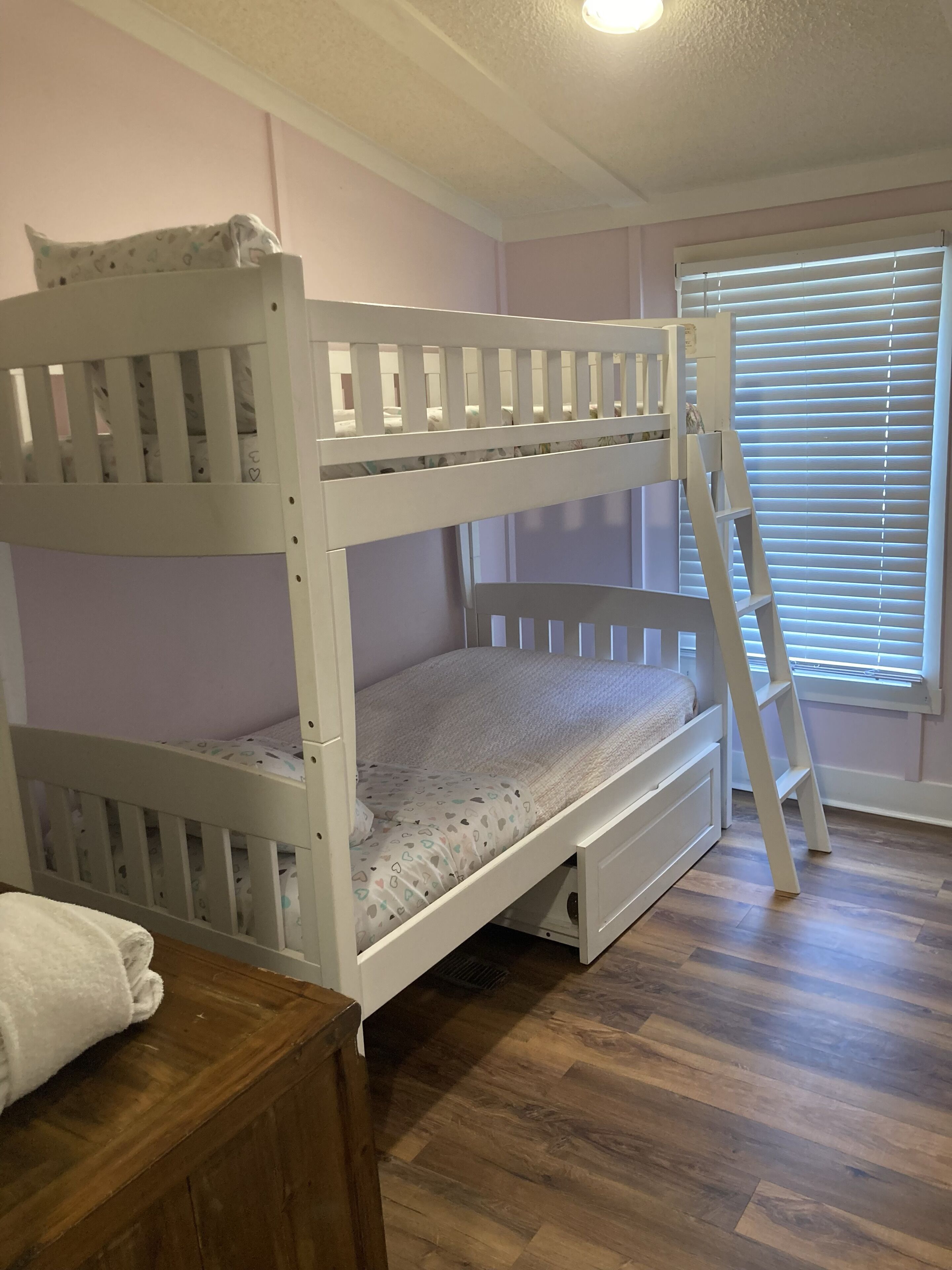 A bunk bed room with a ladder, a window with blinds, and a wooden floor.