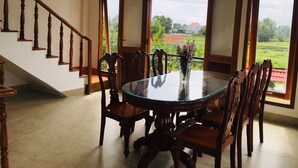 A dining area with a round glass table and wooden chairs, a staircase with wooden balusters, and large windows with a view of greenery and buildings. - Lagi 1001 Nights Hotel (La Gi)