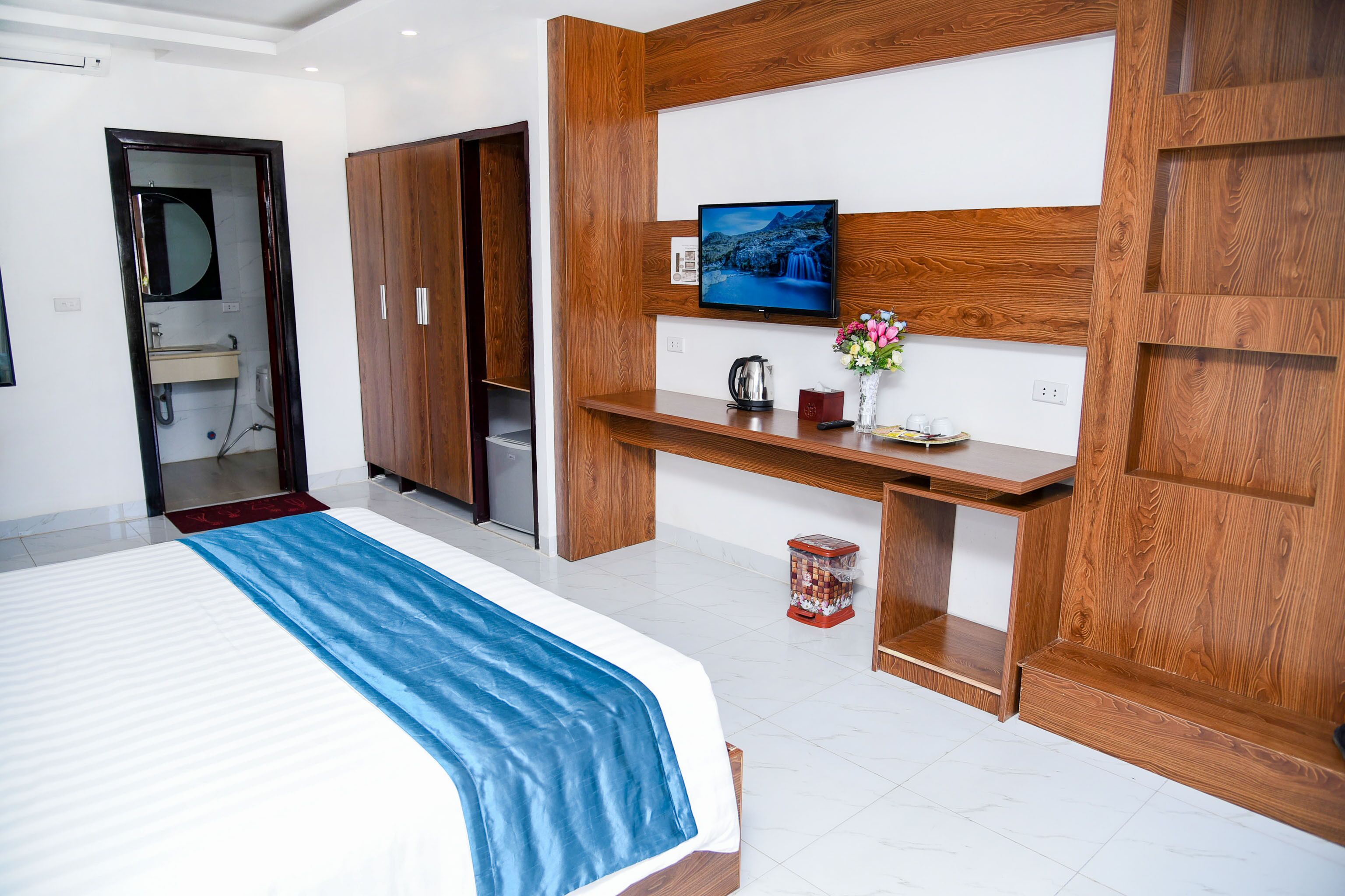 A modern bedroom with a bed, wooden wardrobe, a TV mounted on a wooden panel, and a small shelf with a kettle and flowers.