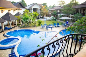 Una piscina con baldosas azules y blancas, rodeada de sillones y mesas, con vistas a casas y vegetación al fondo.