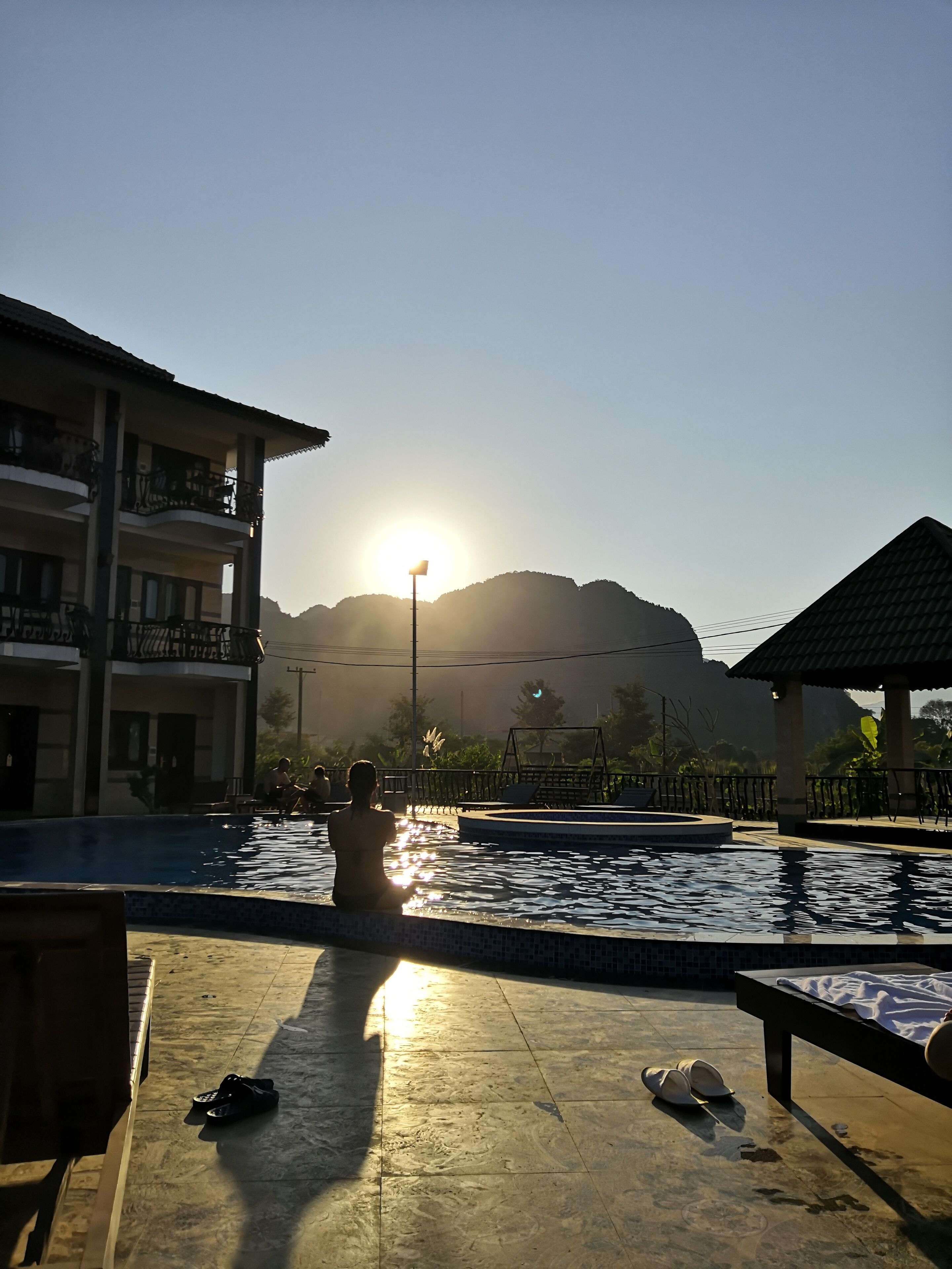 A pool area with a view of mountains, a building with balconies, and a sunlit sky.