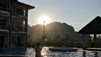 A pool area with a view of mountains, a building with balconies, and a sunlit sky.