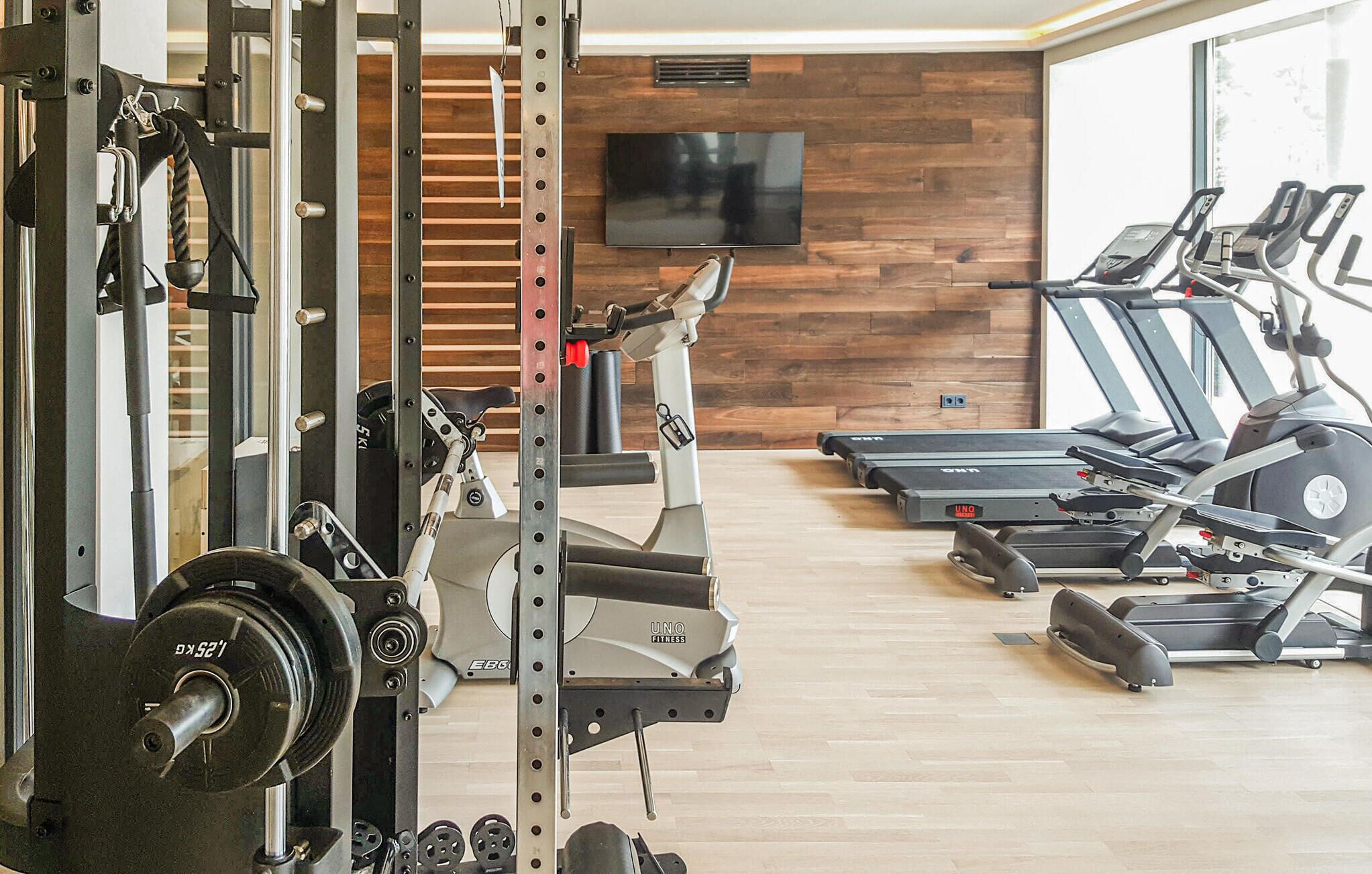 Uma academia com equipamentos de musculação, esteiras e uma TV na parede.