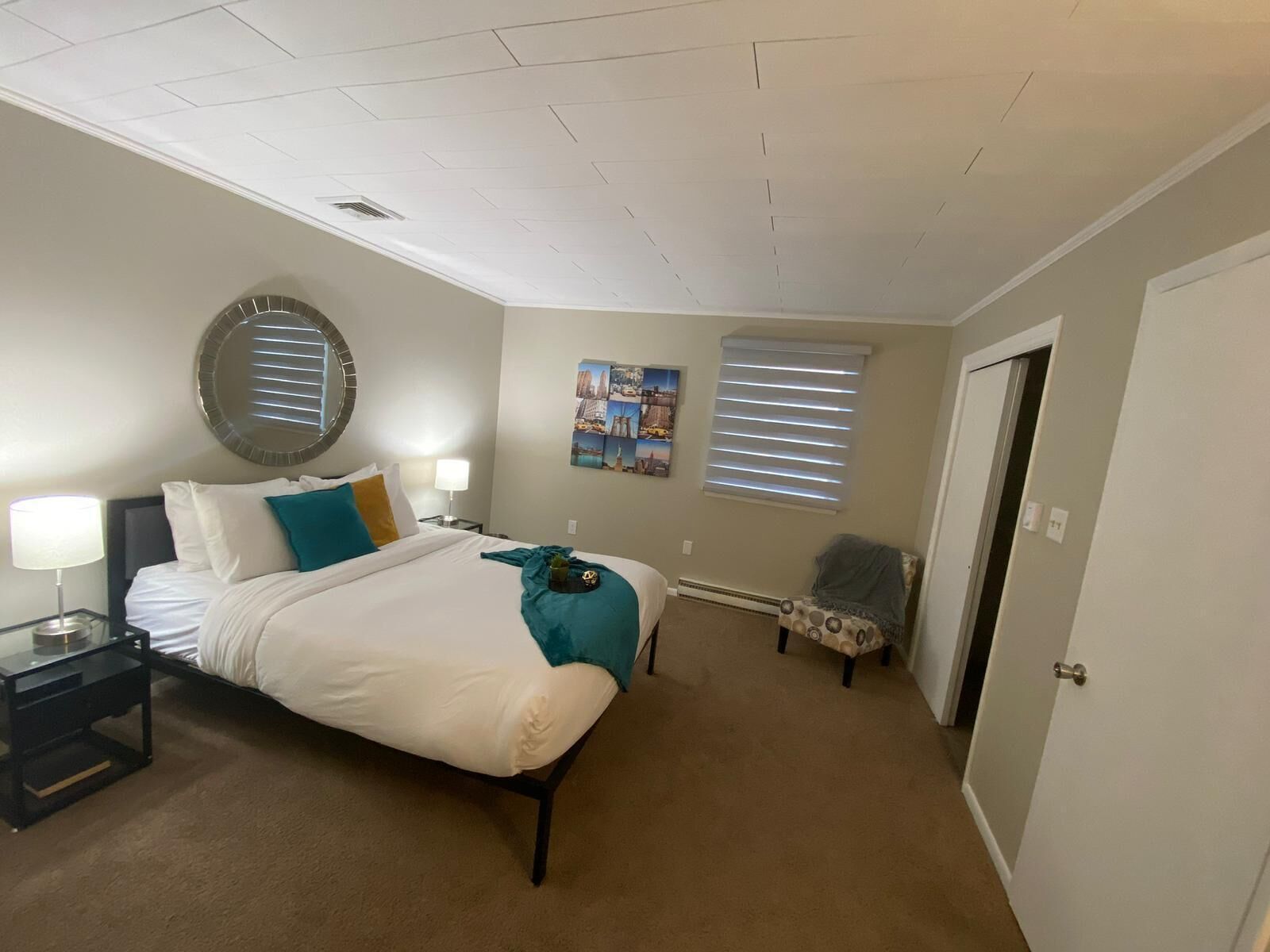 A bedroom with a bed, bedside tables, a mirror, a wall clock, a window with blinds, and a door.