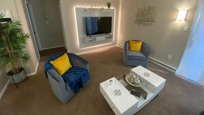 A modern living room with a flat-screen TV mounted on the wall, two armchairs with yellow cushions, a coffee table with decorative items, and a large potted plant. - Close to lot of shopping areas, eateries, bars and more. Ideal for monthly stays (Lancaster)