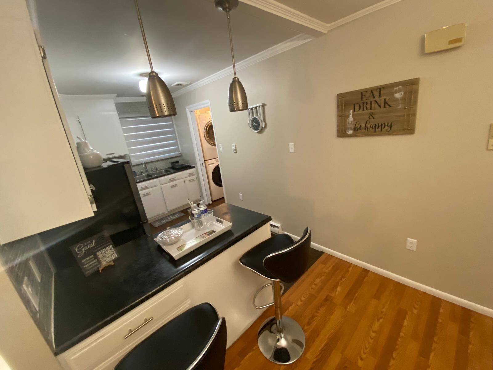 A kitchen with a black countertop, two black bar stools, a clock, and a sign that says "Eat Drink & Be Happy".