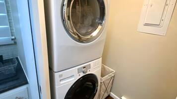 Une petite buanderie comprenant un lave-linge et un sèche-linge superposés, une petite poubelle et une porte blanche.