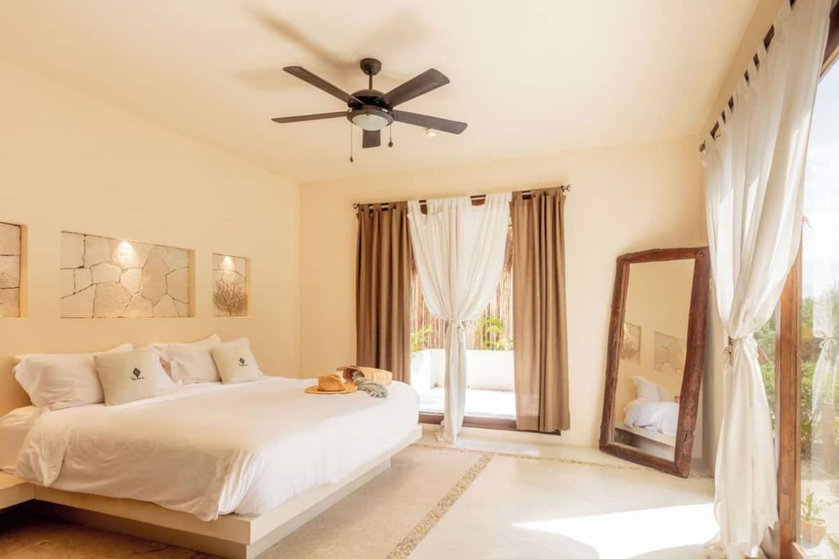 Un dormitorio con cama, ventilador de techo, cortinas y un espejo grande.