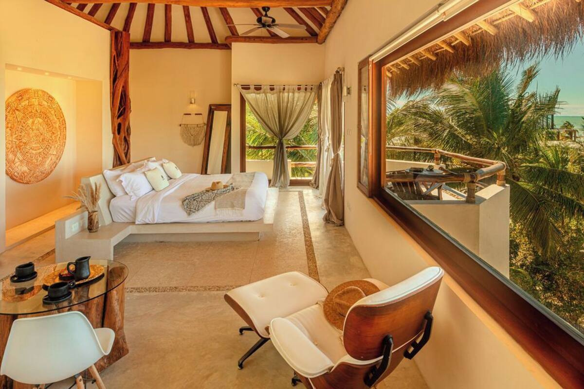Un dormitorio con cama, escritorio, sillas y un gran ventanal con vistas a palmeras y al océano.