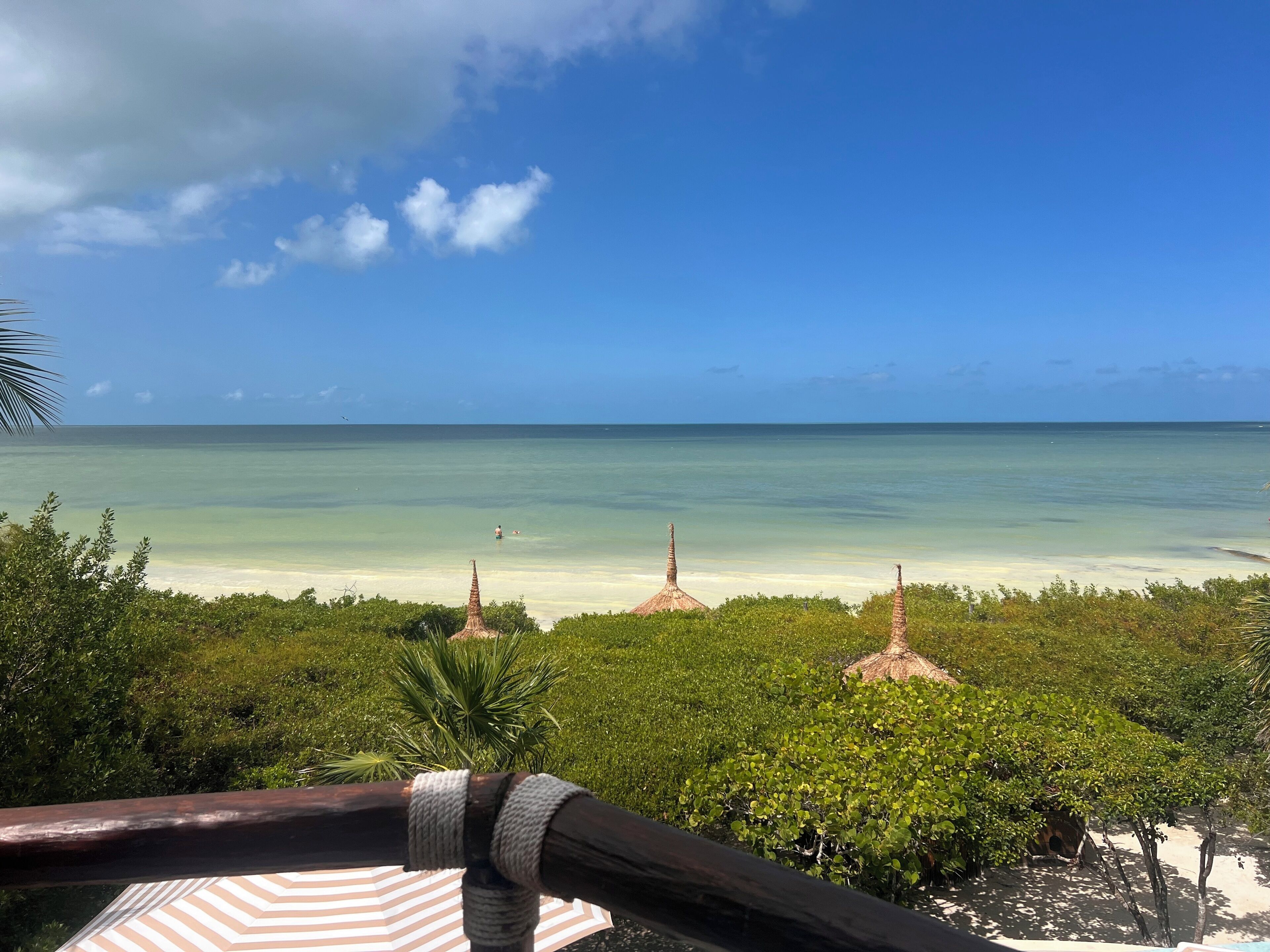 A beach view with clear skies, a calm sea, and thatched huts in the foreground.