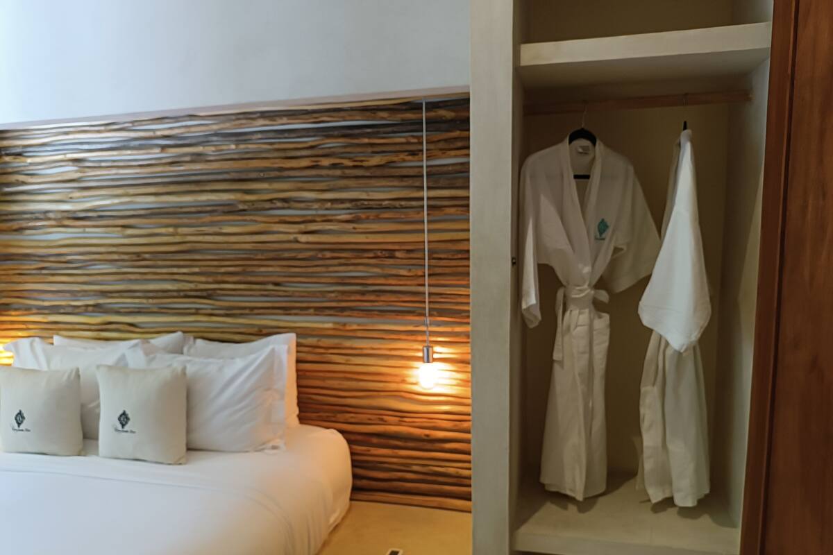 Habitación de hotel con cabecera de madera, cama con ropa de cama blanca, mesita de noche con un control remoto y una bata colgada en un estante.