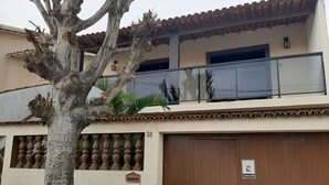 Un immeuble avec une porte marron, un balcon doté d’une rambarde en verre et un arbre devant.