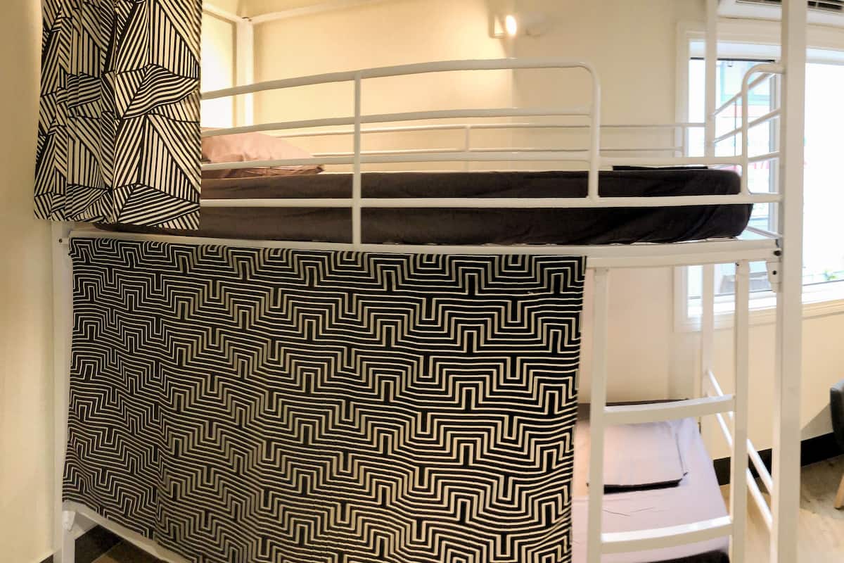 Una cama litera con cubrecama estampado, dos sillas, una mesita y un mueble para guardar objetos en una habitación con ventana.