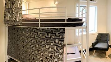 A bunk bed with a patterned bedspread, two chairs, a small table, and a storage unit in a room with a window.