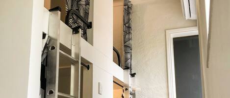 A narrow hallway with a metal ladder, a window with a blind, and a door with a zebra-striped curtain.