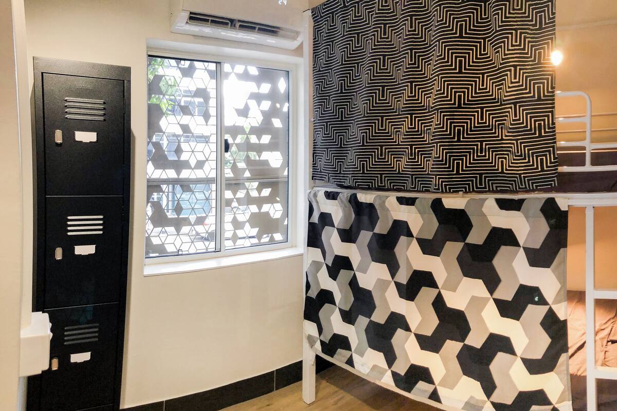 Una cama individual superpuesta con ropa de cama de diseño geométrico, una ventana con mosquitera geométrica y un casillero de metal negro.