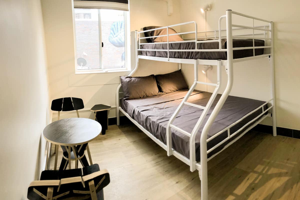 Una cama litera de dos plazas, una pequeña mesa con sillas, una lámpara y una ventana con persianas.