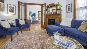 Un salon avec un canapé bleu, deux fauteuils bleus, un tapis à motifs, une cheminée et une bibliothèque.