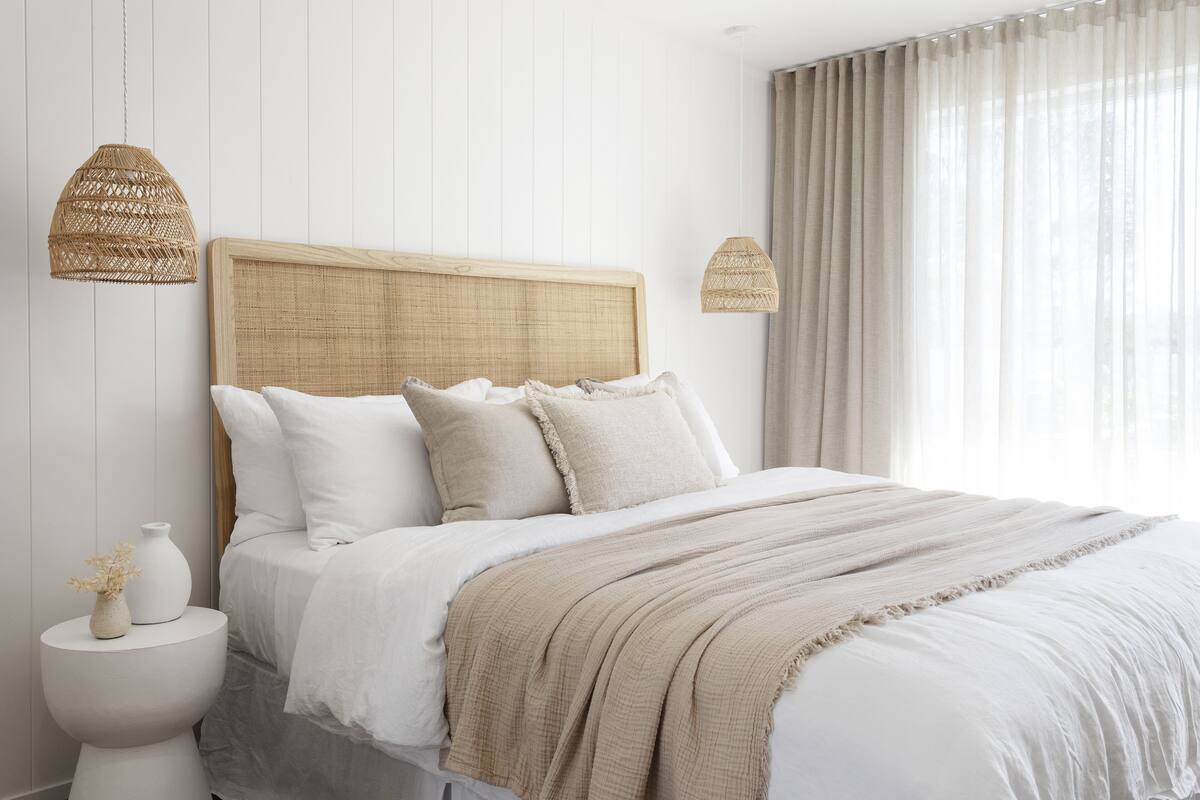 Une chambre avec une tête de lit en bois, du linge de lit blanc, un plaid beige et deux suspensions lumineuses en osier.