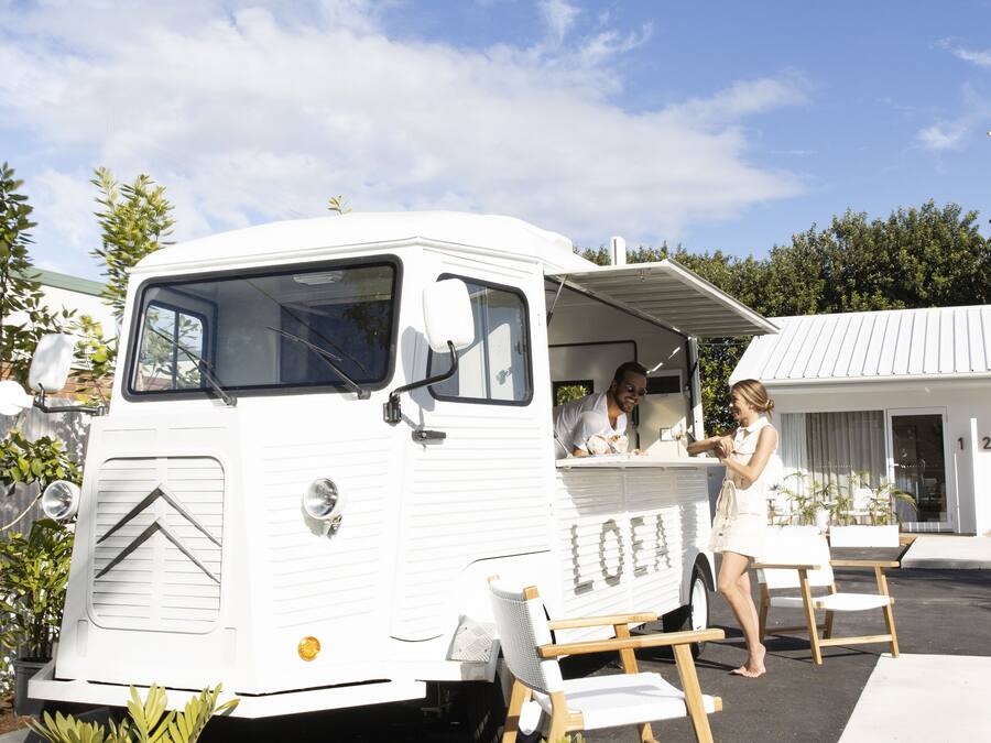 Un camión de comida de estilo vintage en blanco con la palabra "LOEA" en su lateral, estacionado frente a un edificio moderno con fachada blanca y el número "12" a la vista.