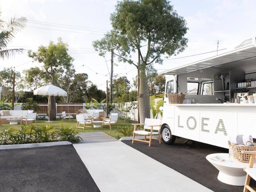 Un camión de comida blanco con la marca "LOEA" estacionado en un área exterior bien cuidada, con mesas y plantas.