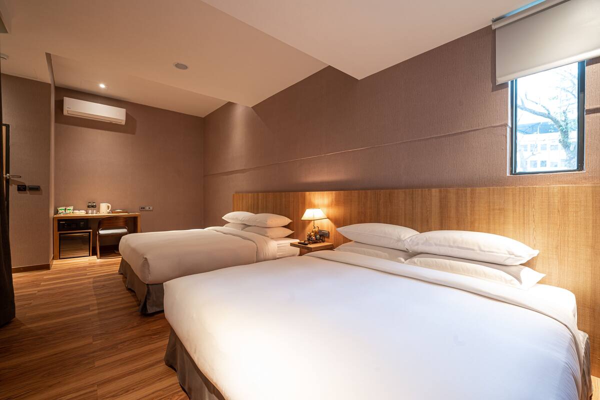 A hotel room with two beds, a desk, a lamp, and a window.