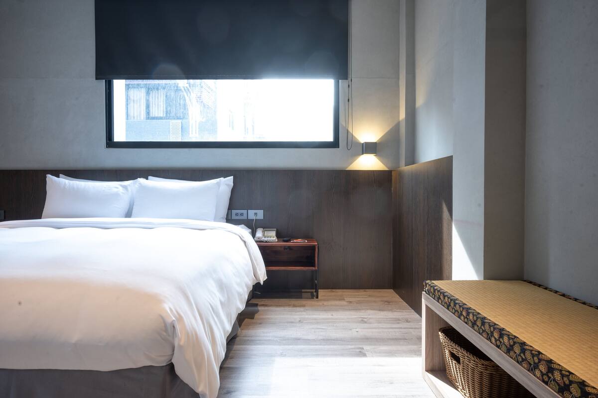 A modern hotel room with a large bed, a bedside table with a lamp, a small table with a basket, and a window with a view of buildings.