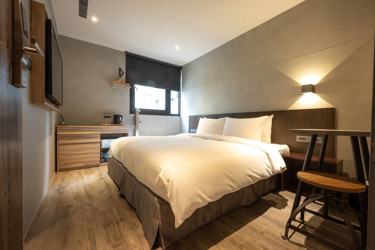 A modern bedroom with a large bed, wooden floor, a desk, and a hanging light fixture.