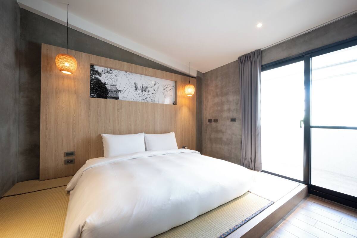 A modern hotel room with a large bed, wooden headboard, and a large window with curtains.
