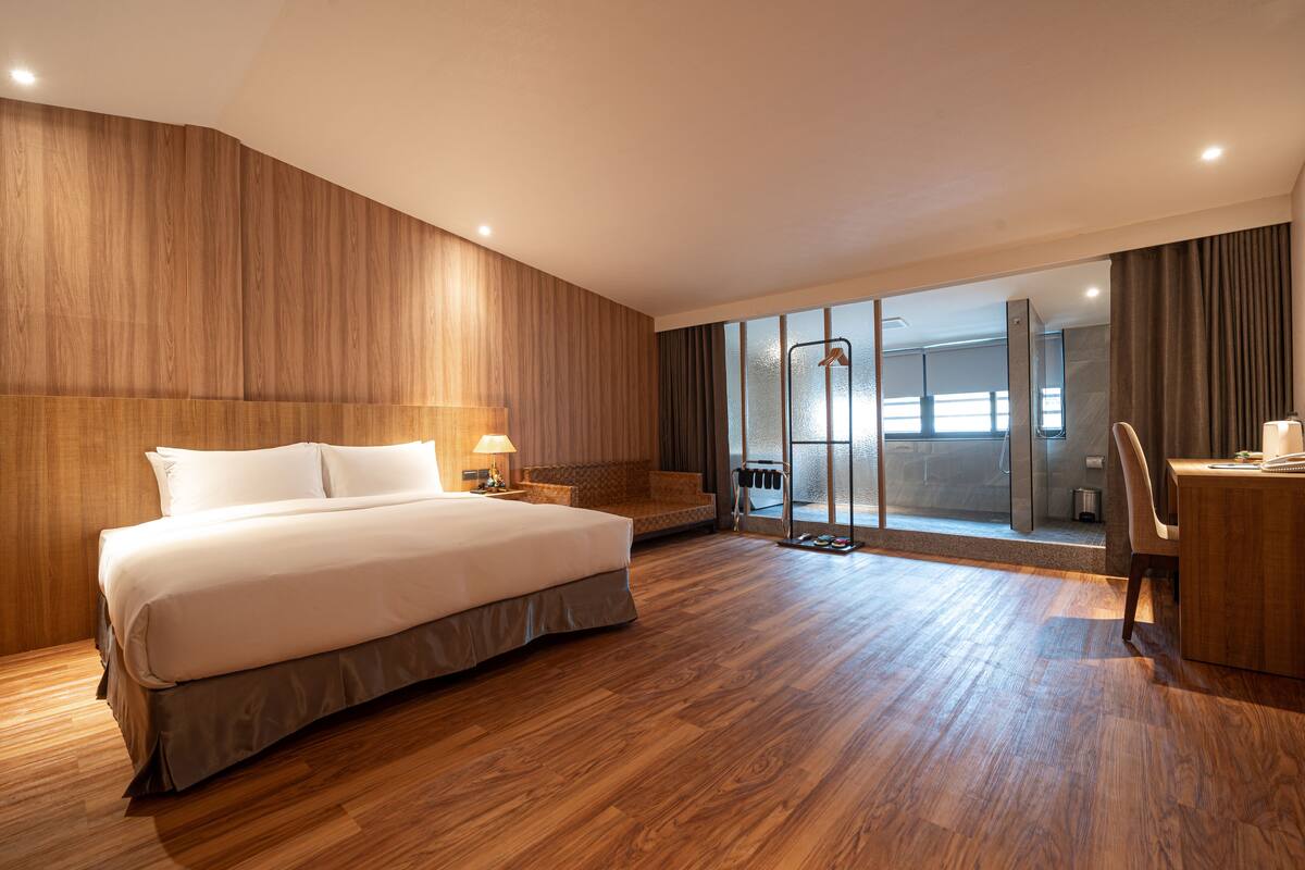 A hotel room with a large bed, a desk, and a view of the city through a glass door.